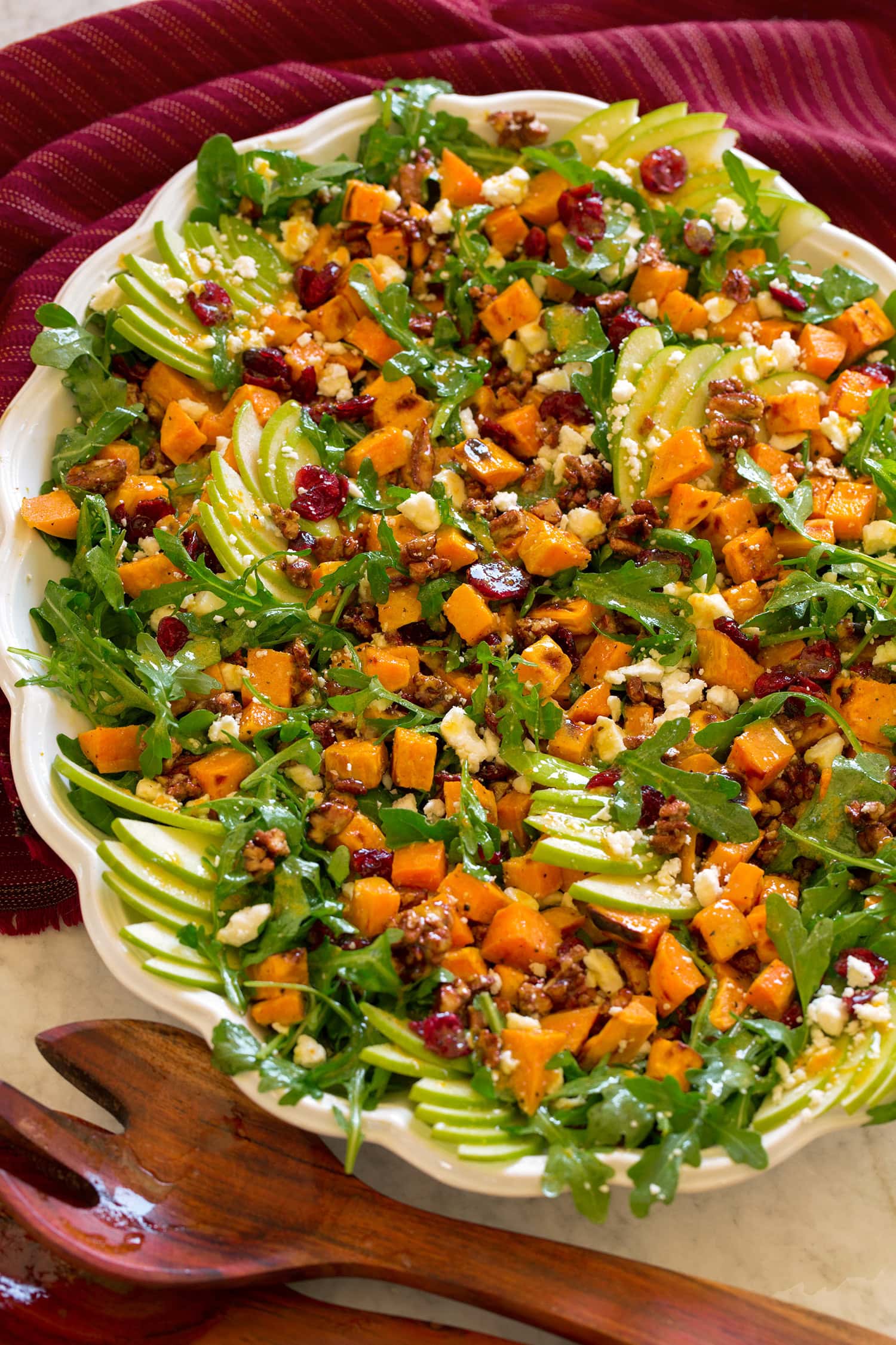 Saldžiųjų bulvių salotos, pavaizduotos iš šono ant baltos ovalios lėkštės su medinėmis žnyplėmis į šoną.