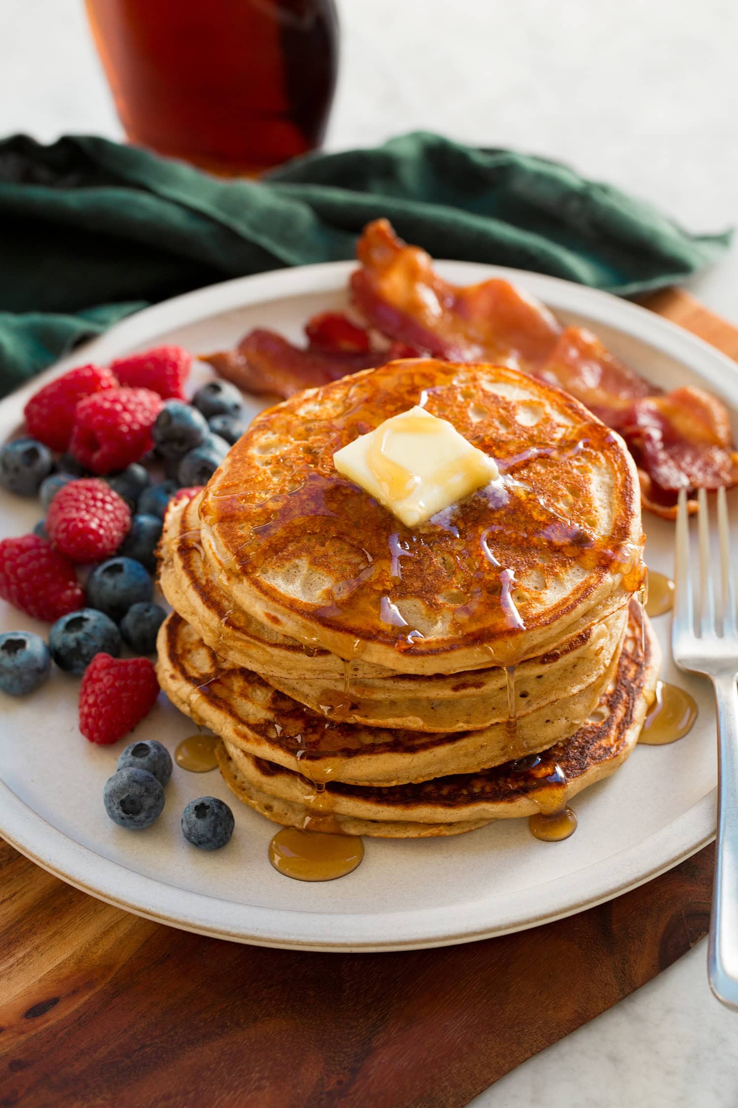 Protein pancakes topped with butter and syrup, shown served with berries and bacon on the side.