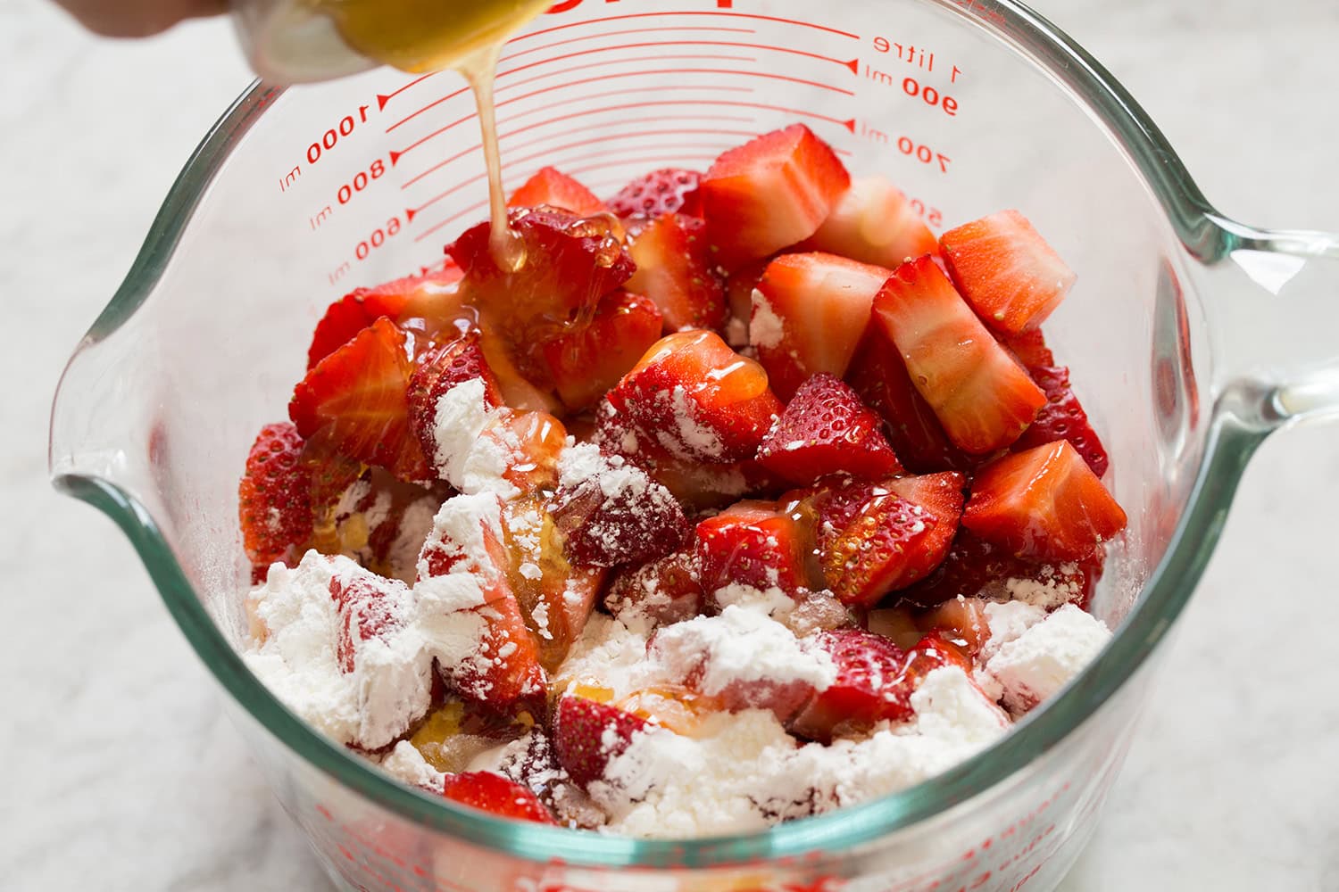 Diced strawberries and cornstarch in a large liquid measuring cup with honey being drizzled in.