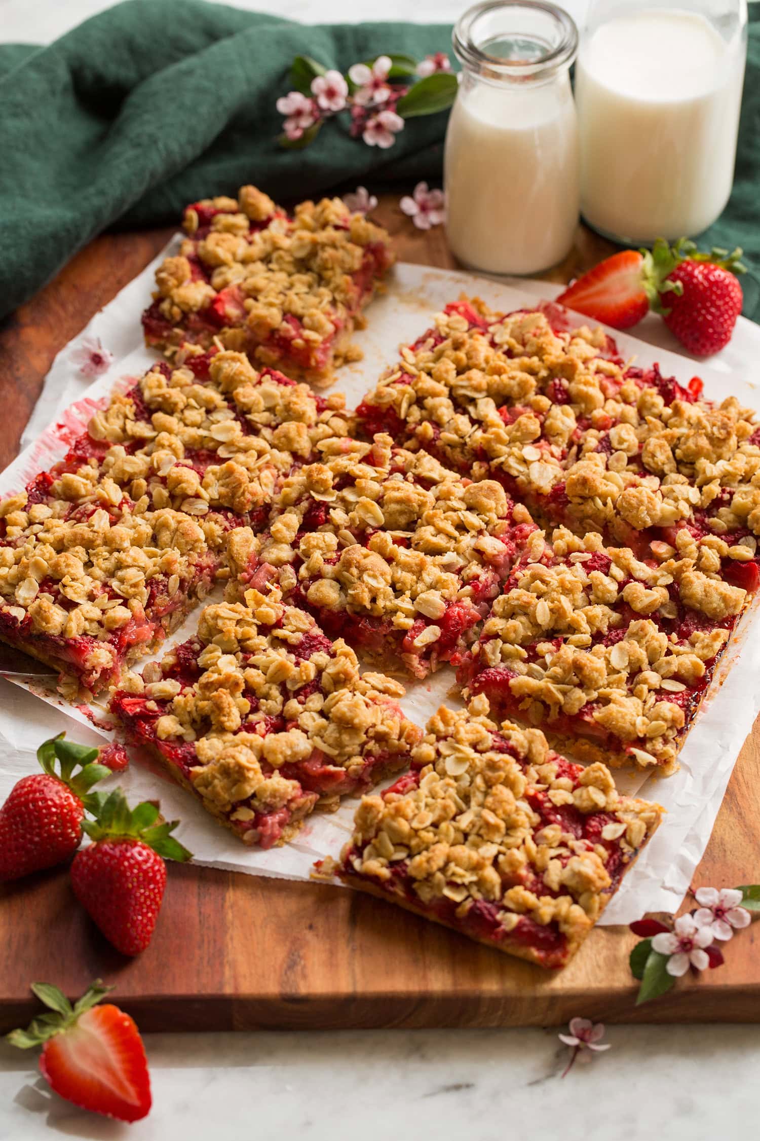 Strawberry filled oatmeal bars shown on white parchment set over a wooden serving tray. They are cut to show filling and serving size. Decorative fresh strawberries are shown to the sides.