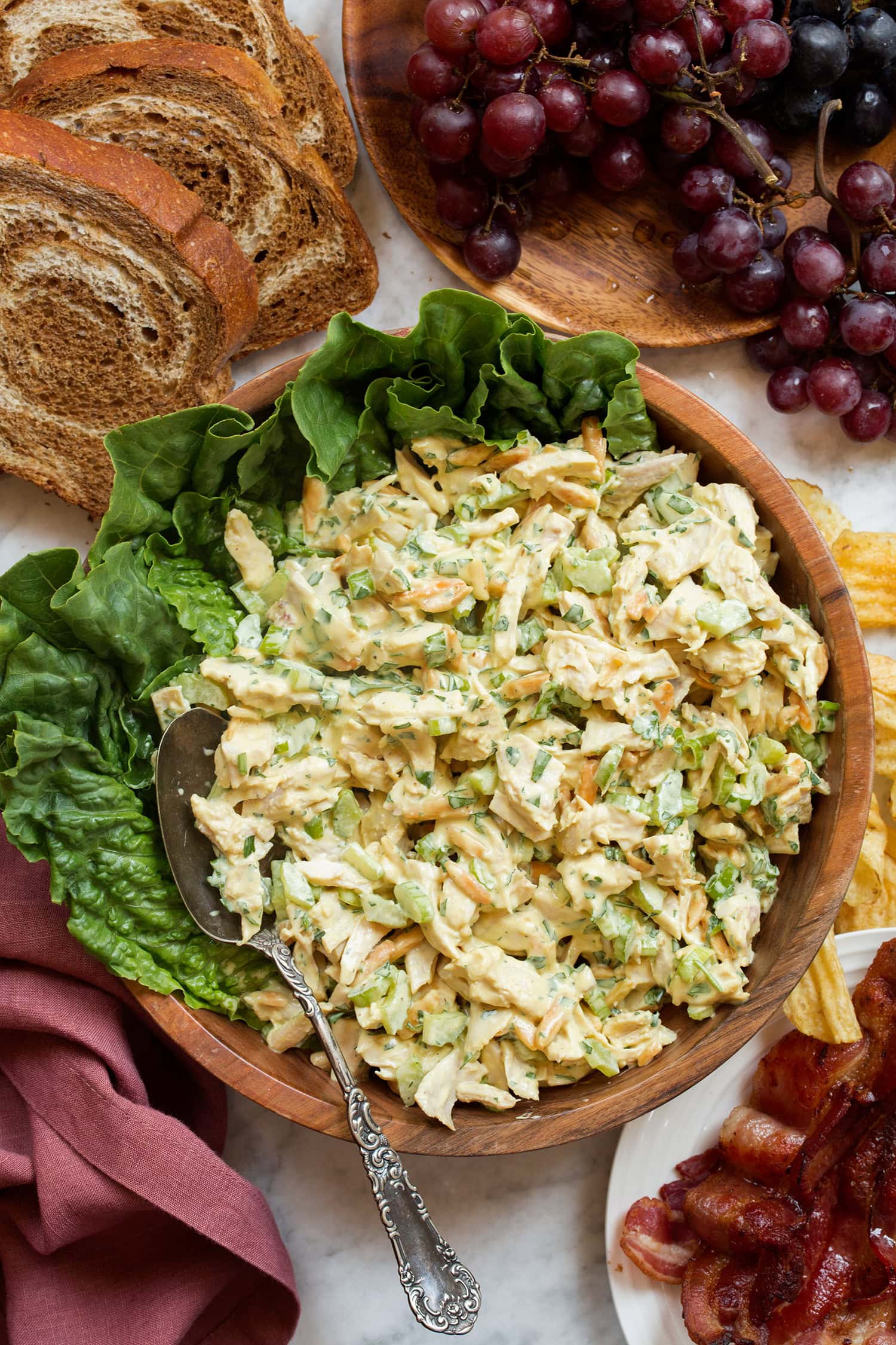 Bowl of chicken salad with honey mustard sauce, shown in a wooden bowl with lettuce leaves. Grapes, bacon and bread slices are laid decoratively around the bowl.