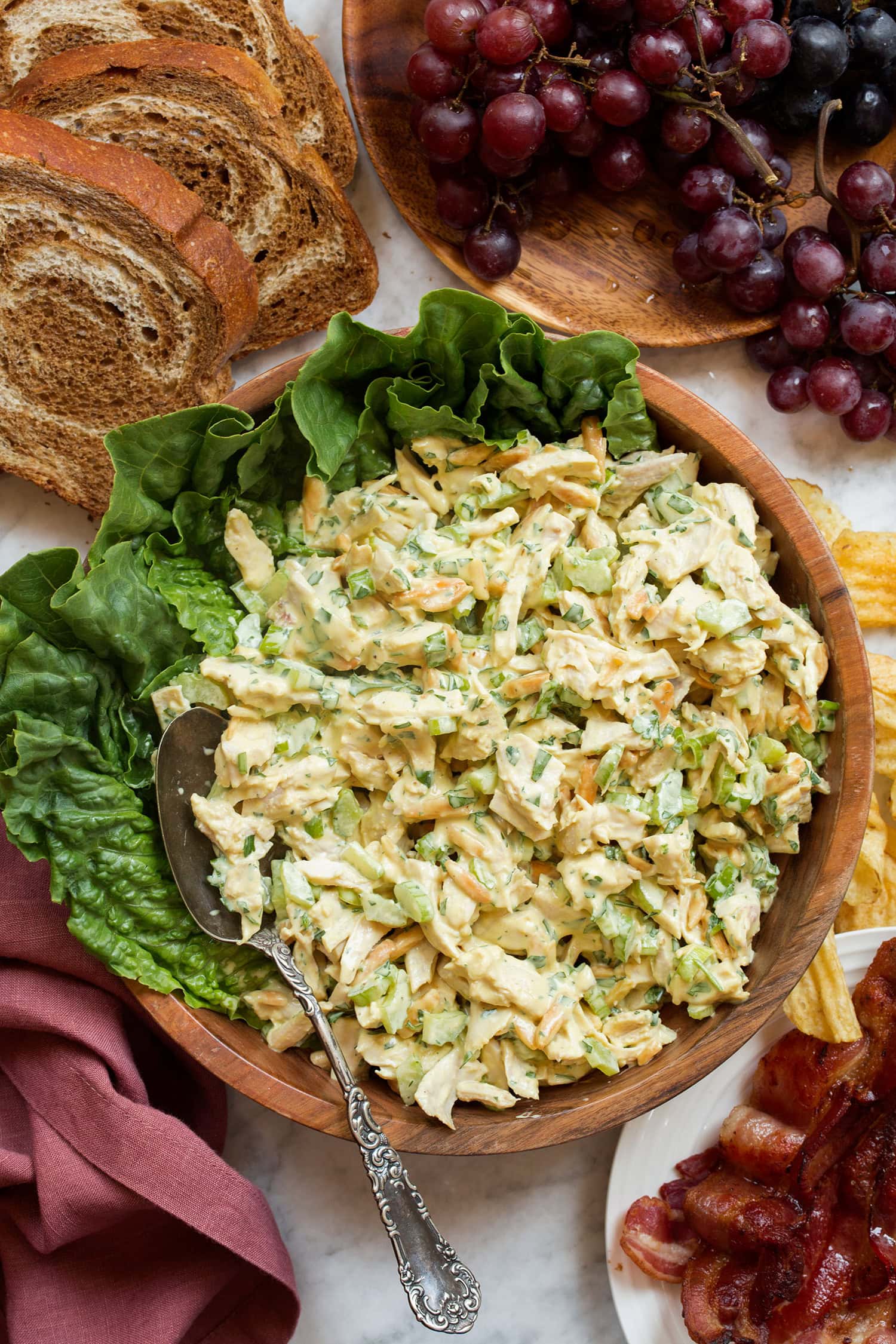 Bowl of of chicken salad with honey mustard sauce, shown in a wooden bowl with lettuce leaves. Grapes, bacon and bread slices are laid decoratively around the bowl.